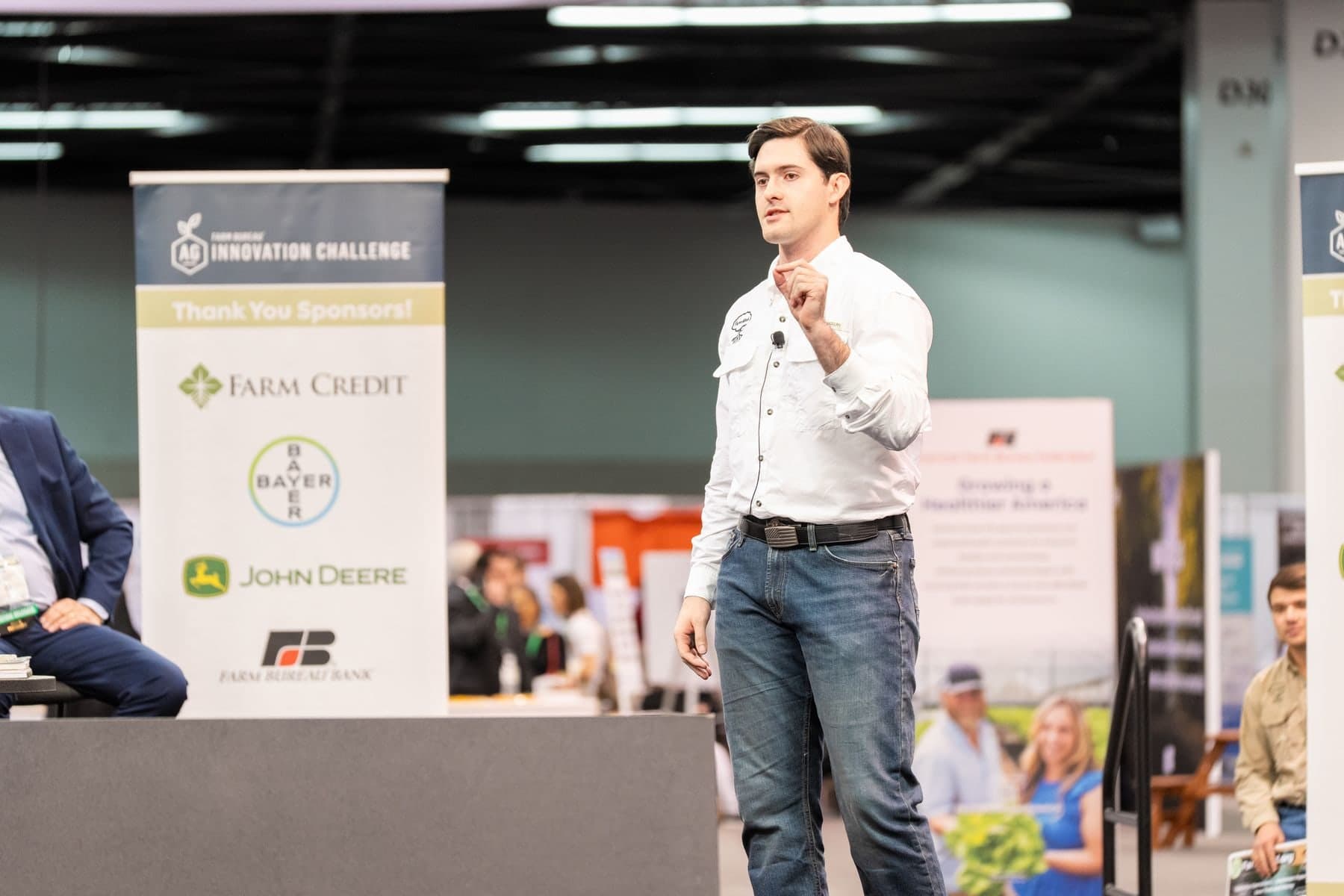 Colin Raby, in a white FarmMind shirt and jeans, pitching on stage at the Farm Bureau Ag Innovation Challenge, with sponsor banners behind him