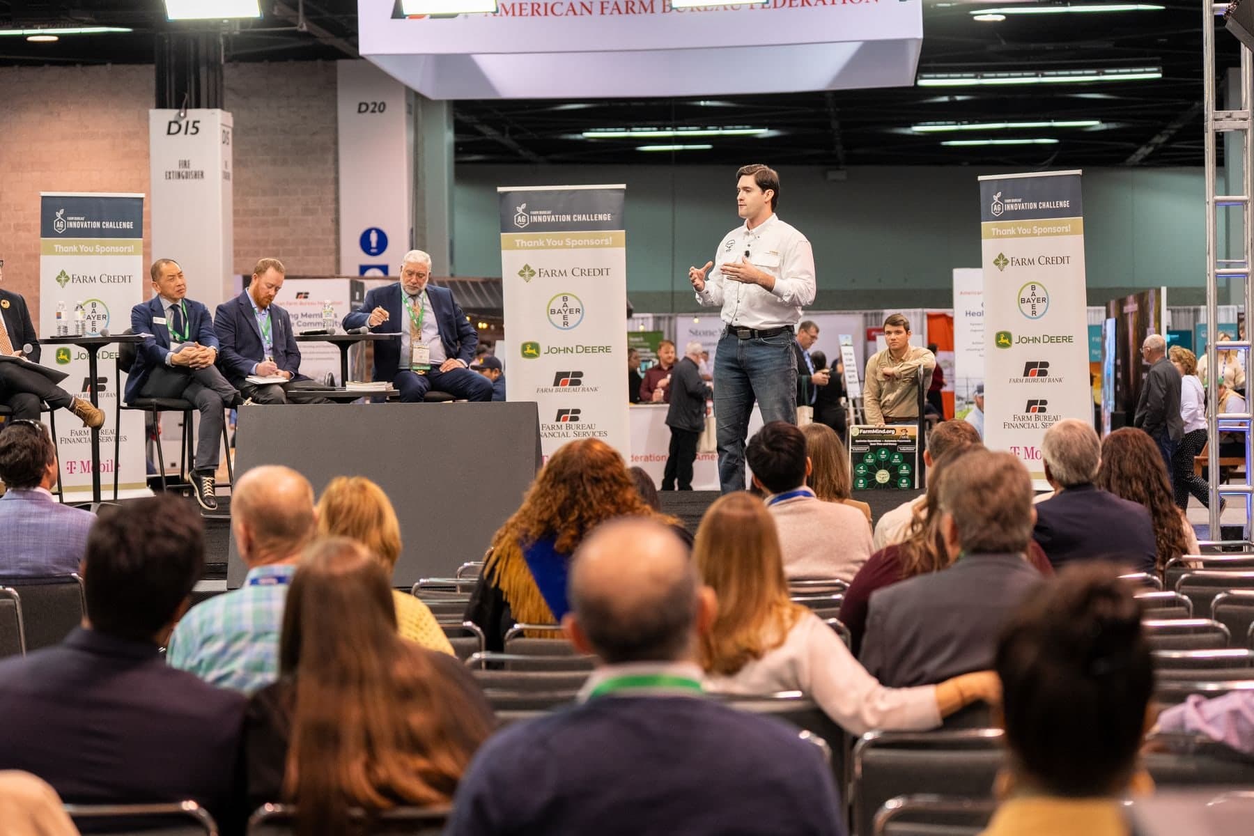 Colin Raby presenting on the Ag Innovation Challenge stage to a seated audience while three judges look on from the left