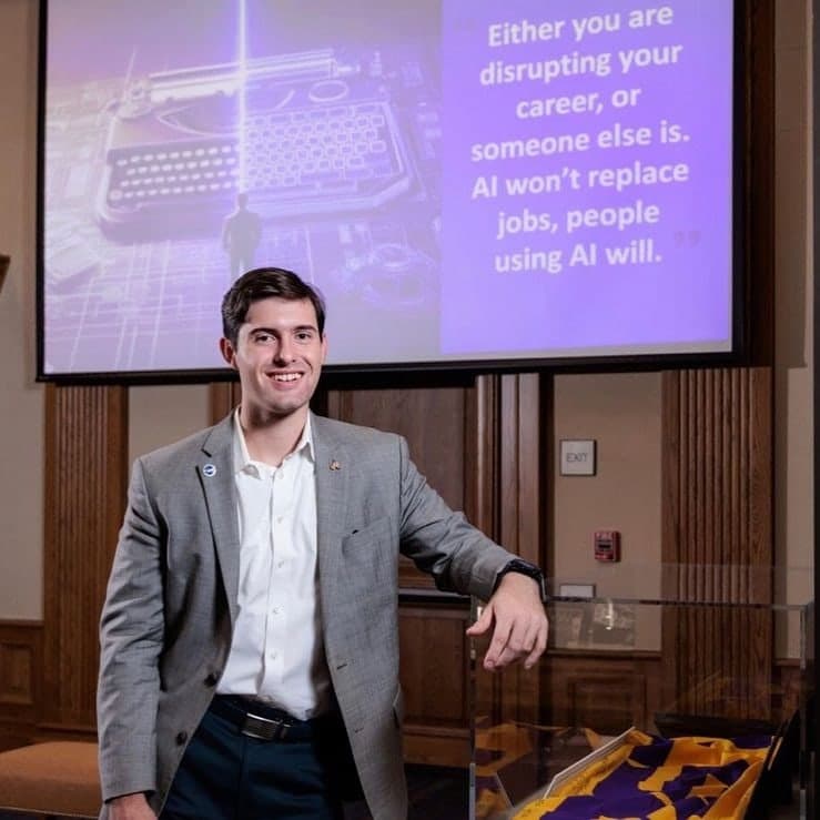 Colin Raby in a gray blazer leaning on a display case beneath a screen reading “Either you are disrupting your career, or someone else is. AI won't replace jobs, people using AI will.”