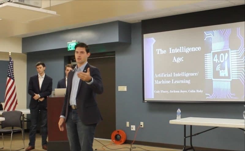 Colin Raby presenting "The Intelligence Age" to the St. Charles Community Advisory Panel, his title slide on the screen behind him and teammates Cody Flurry and Jackson Joyce at left