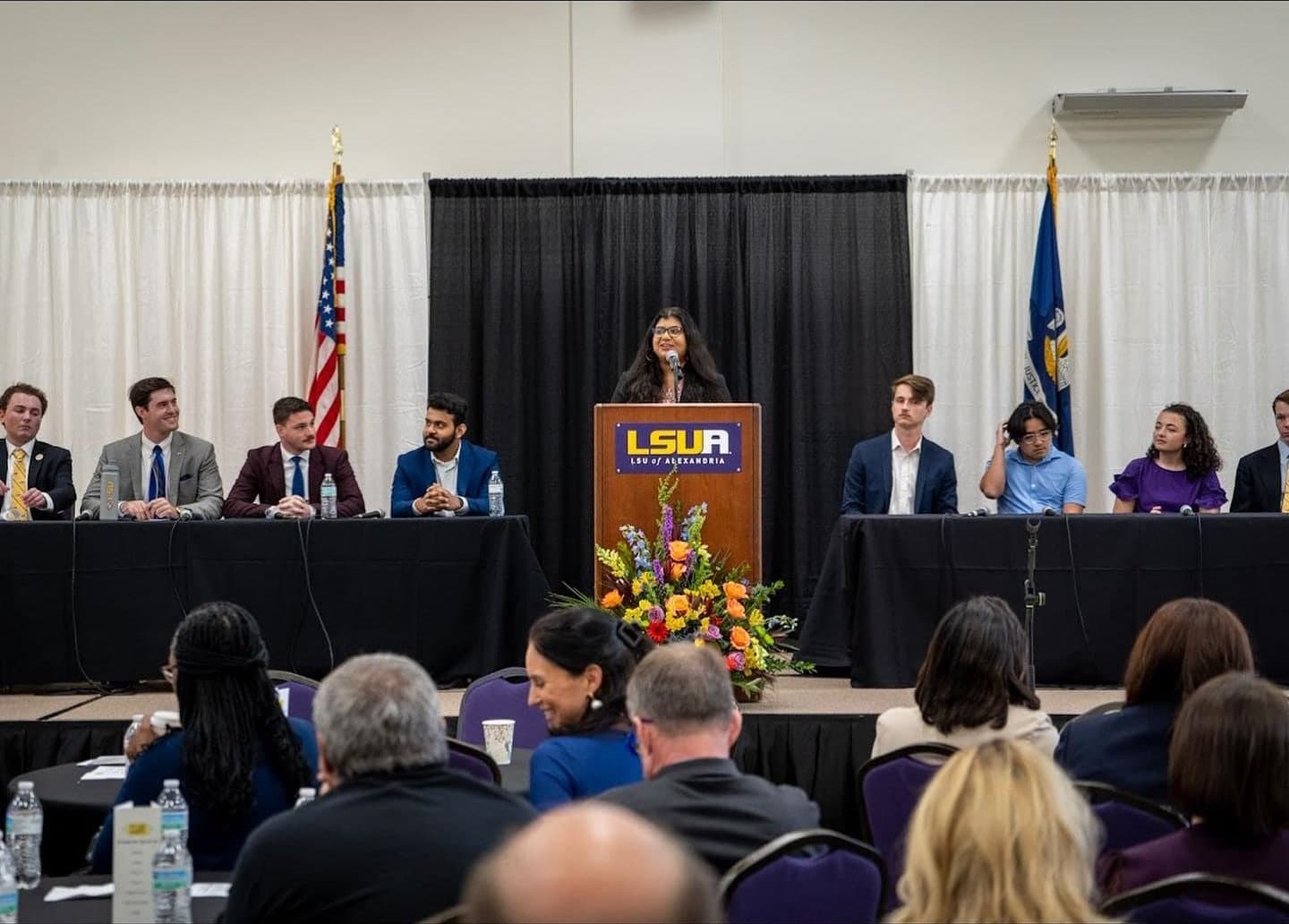 A student panel seated at two long tables on either side of an LSU of Alexandria podium, with the American and Louisiana flags behind them; Colin Raby, in a gray suit, is second from left