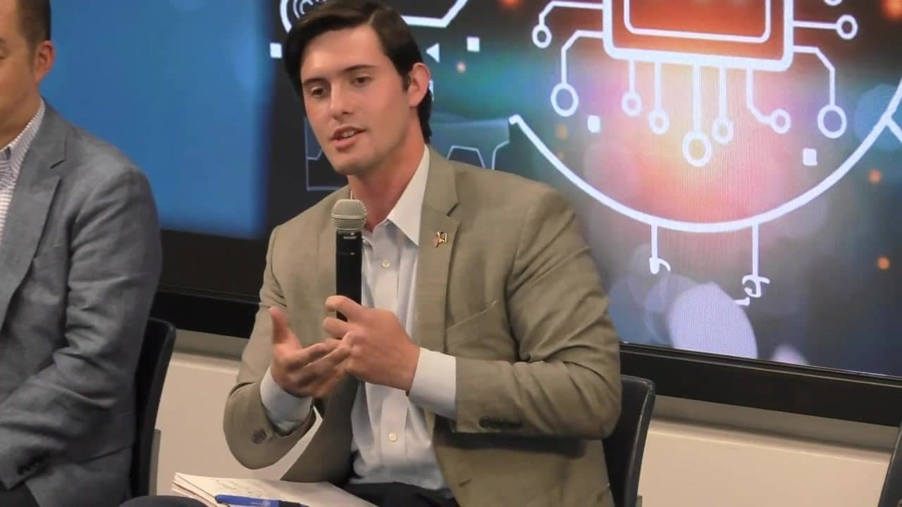 Colin Raby speaking into a microphone on the panel, an AI circuit graphic on the screen behind him