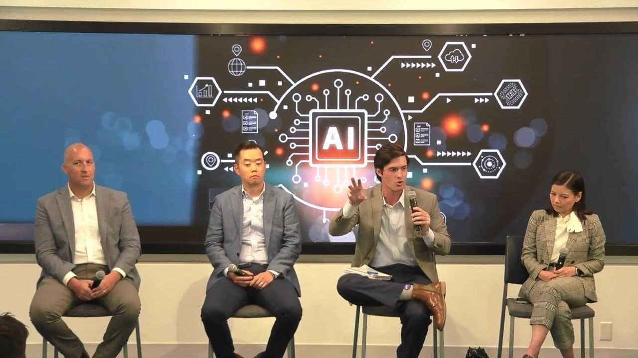 The four panelists seated in front of a large AI graphic, Colin Raby third from left holding the microphone