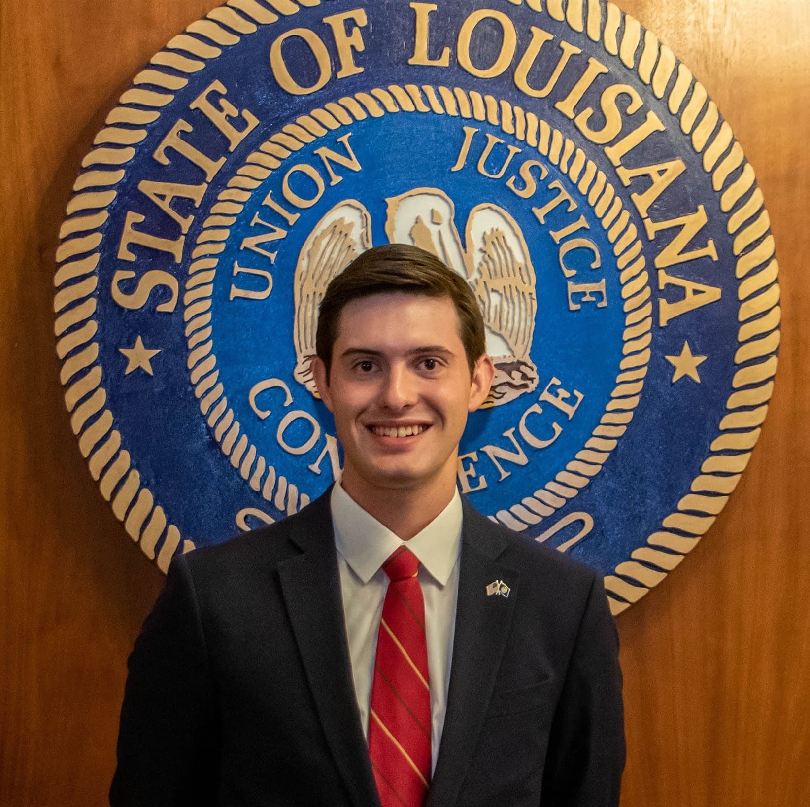 Colin Raby in a suit and red tie in front of the Great Seal of the State of Louisiana