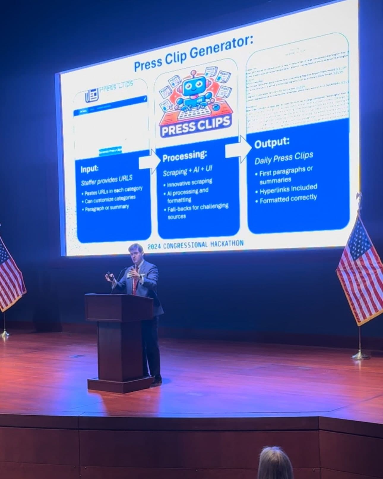 Colin Raby at the podium beneath a slide diagramming the Press Clips Generator's inputs, processing and outputs