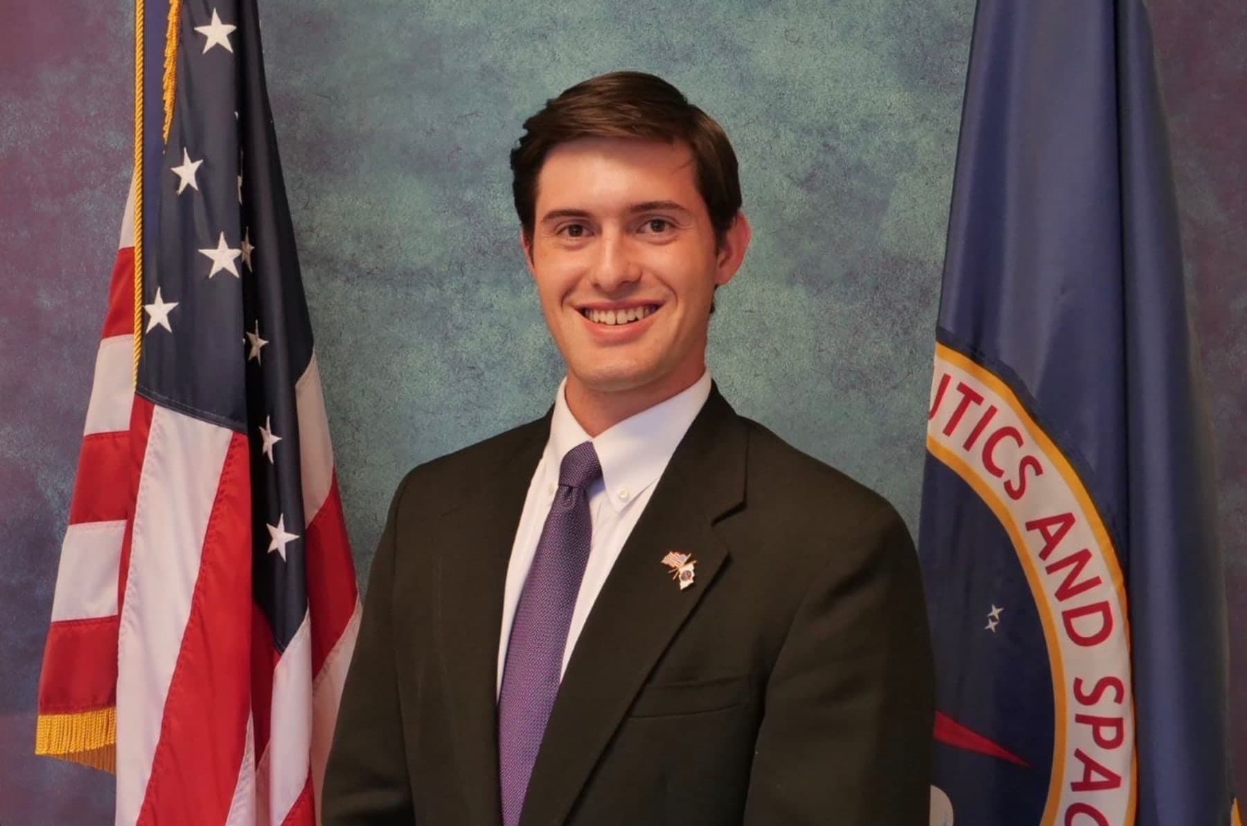 Official portrait of Colin Raby in a dark suit and purple tie, between the American flag and the NASA flag