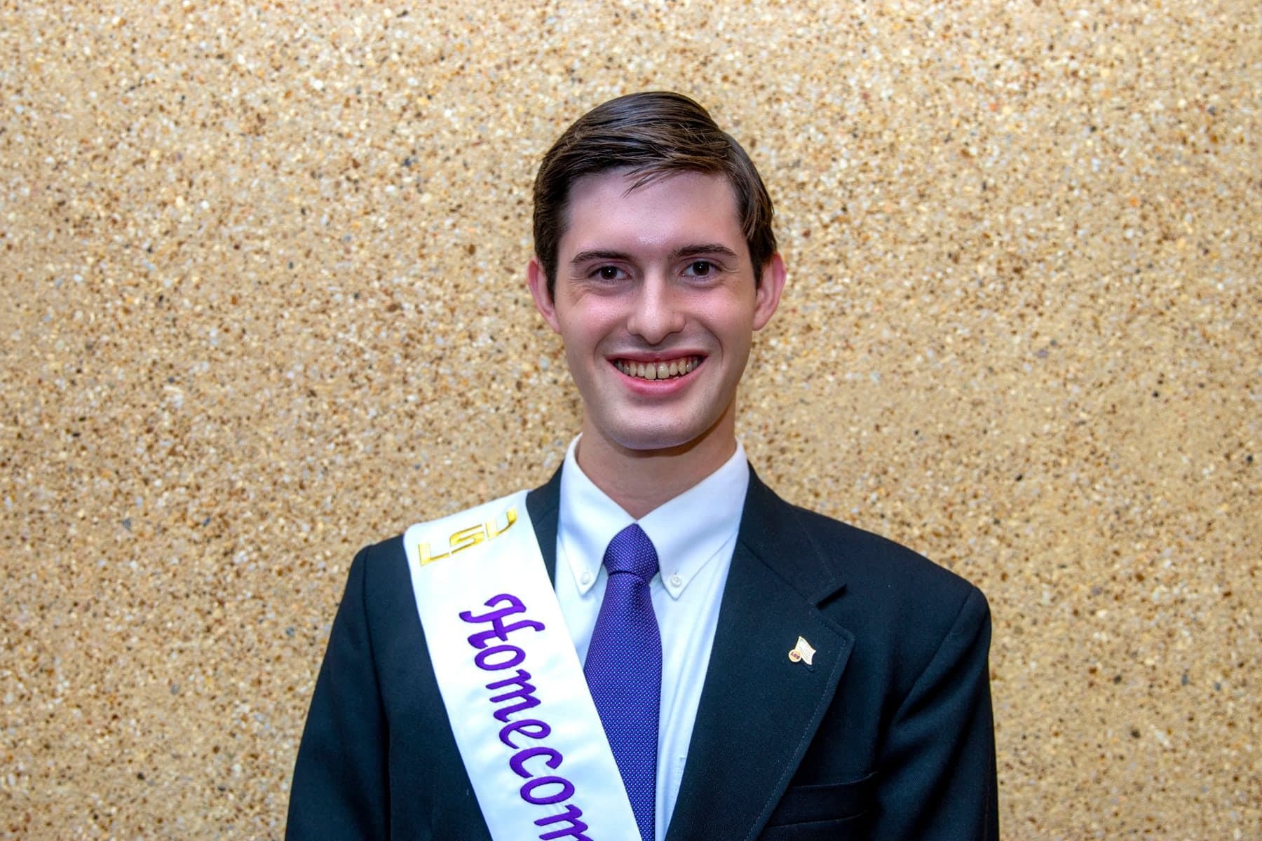 Colin Raby in a navy suit and purple tie wearing an LSU Homecoming sash