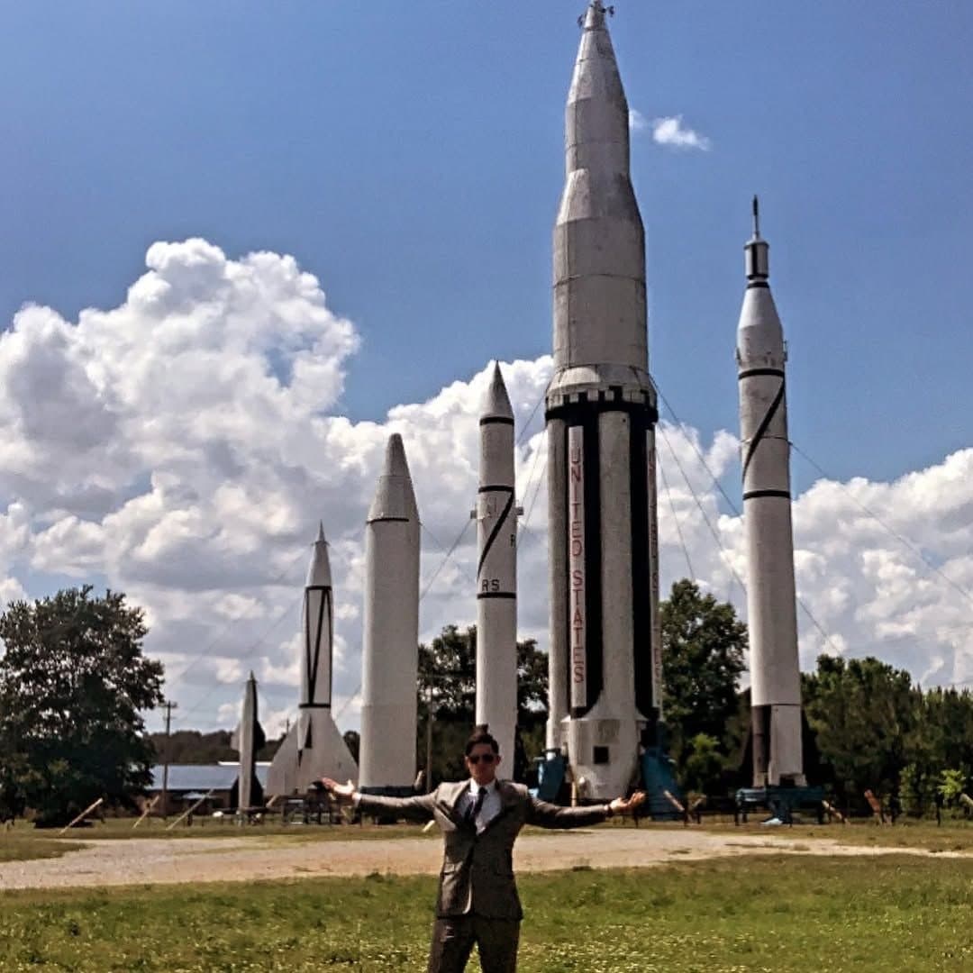 Colin Raby with arms outstretched beneath a row of historic rockets in Huntsville, Alabama