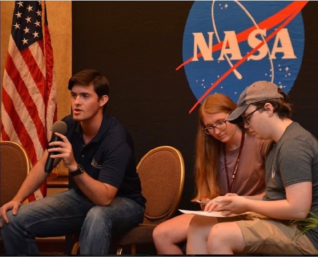 Colin Raby speaking into a microphone on a panel with fellow NASA interns in front of the NASA logo