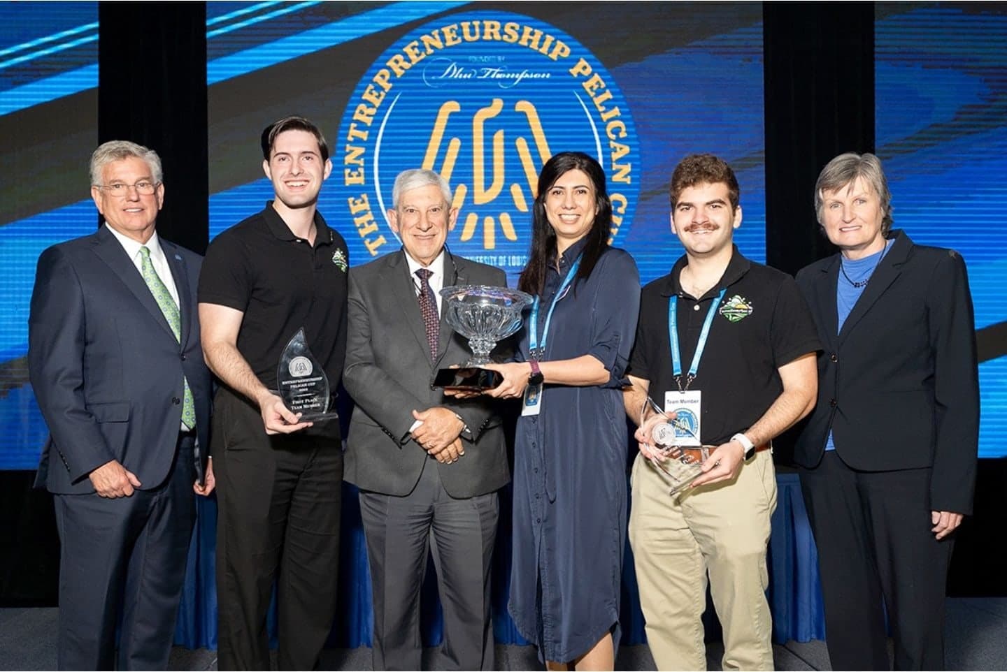 Colin Raby and Grant Muslow holding first-place awards on stage at the Entrepreneurship Pelican Cup with Dhu Thompson, Dr. Nick Bruno, Fahimeh Abbasi and Dr. Michelle McEacharn
