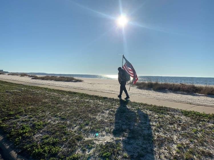A marcher carrying a large American flag along a sunny Gulf Coast beach road, casting a long shadow