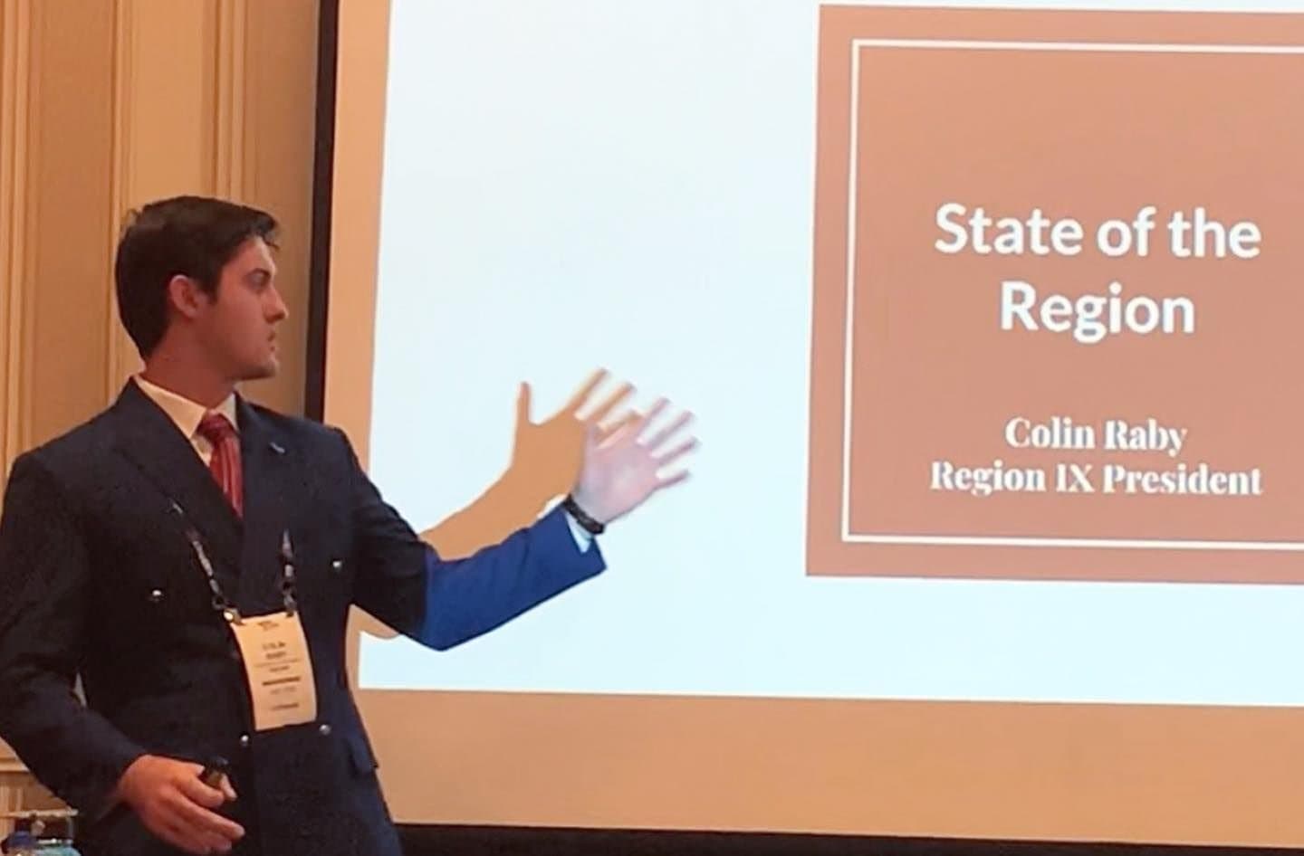Colin Raby in a blue blazer presenting beside a slide reading “State of the Region, Colin Raby, Region IX President”