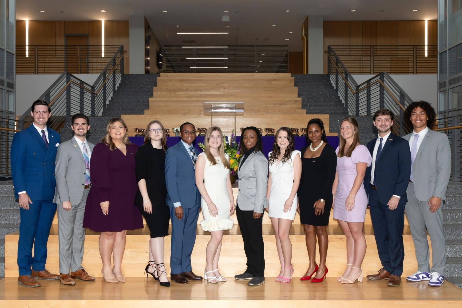 The 12 members of LSU's 2024 Tiger Twelve standing in a row on wooden steps in a campus atrium; Colin Raby, in a blue double-breasted suit, is at far left