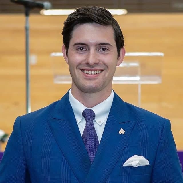 Colin Raby in a blue suit and purple tie, smiling, in a campus atrium