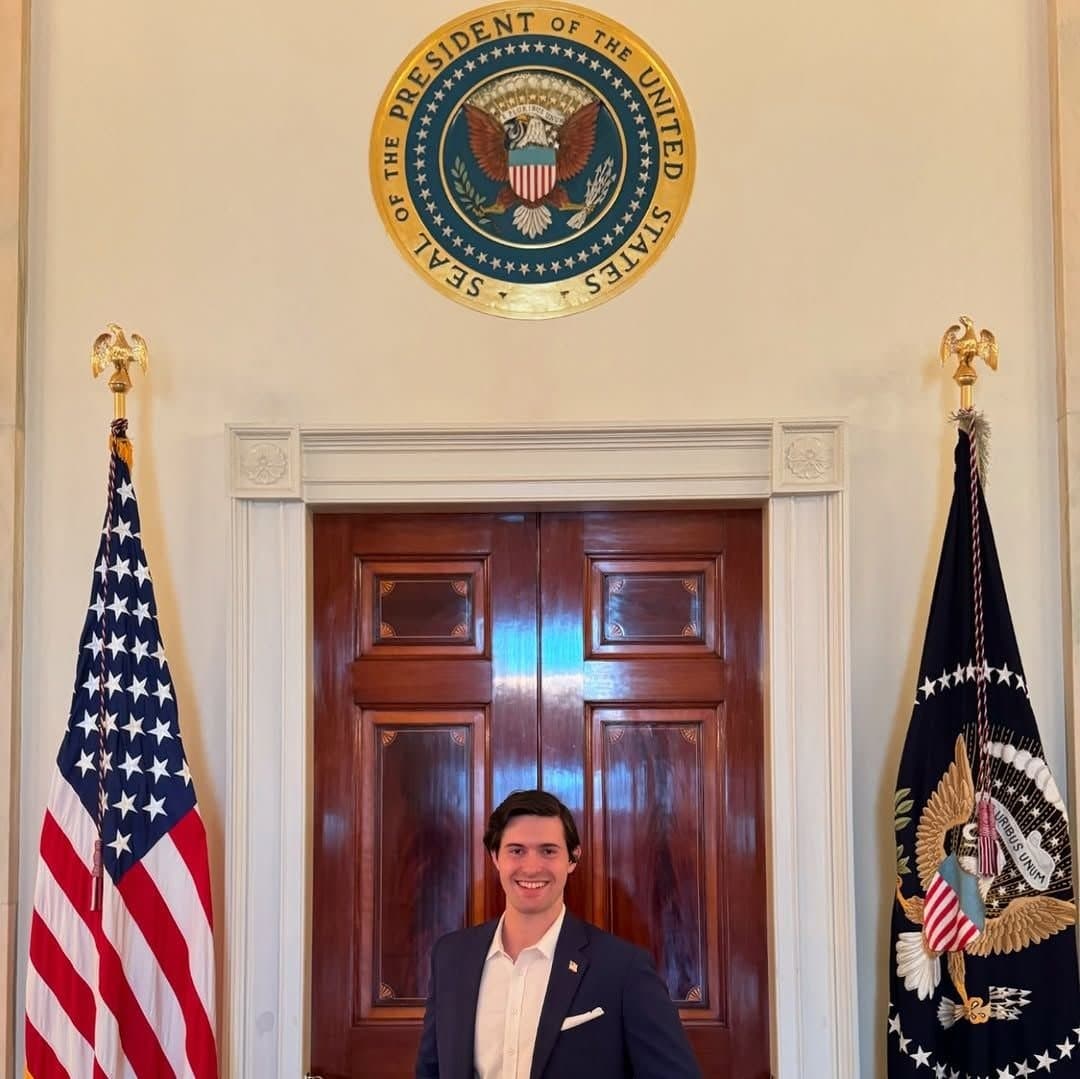 Colin Raby standing in a White House doorway beneath the Seal of the President of the United States, flanked by flags