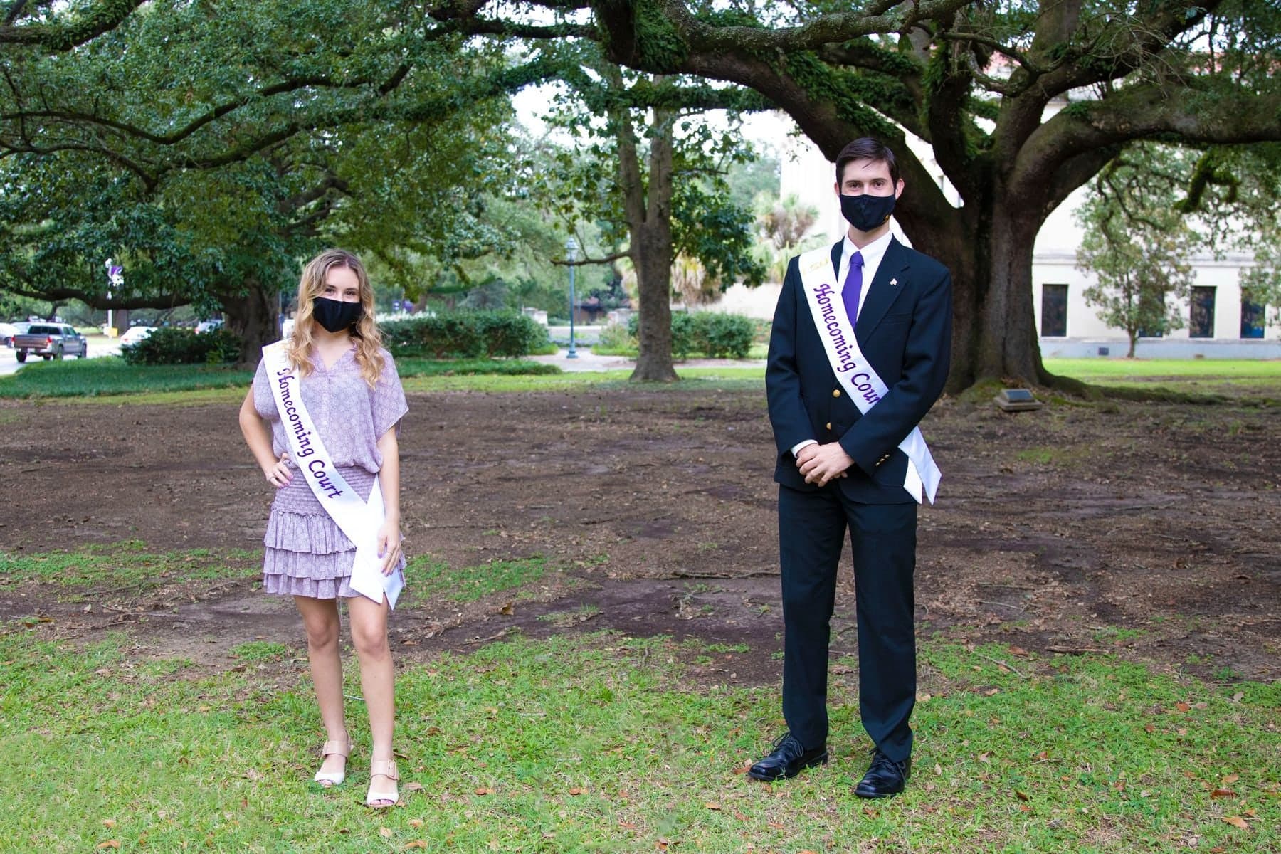 Colin Raby and a fellow court member, both masked and wearing Homecoming sashes, standing under the oaks on campus