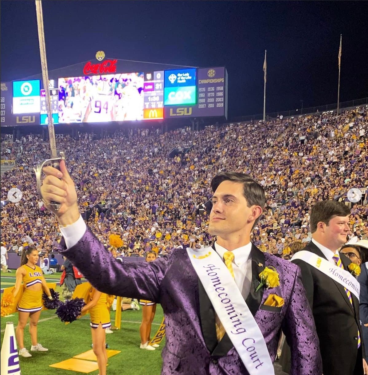 Colin Raby in a purple suit and Homecoming Court sash raising a sword on the field in a packed Tiger Stadium at night